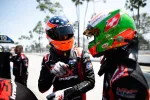 Two race car drivers in colorful helmets and racing suits sharing a moment at the pit stop on a sunny day.