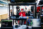 Race team members in helmets and gear sitting in the pit area focused on monitors during a motorsport event.