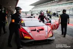 Red race car in pit stop with team members preparing driver for track at a professional racing circuit.
