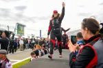 Female race car driver in black and red suit waves to crowd while walking on stage at racetrack event.