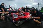 Mechanics inspecting and working on a red race car in a pit lane with overcast sky and racing equipment around.