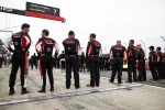 Race team members in black and red uniforms stand in a line on the Porsche Penske Motorsport pit lane before a race.