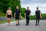 Four people walking on a paved road surrounded by greenery, two men in black shirts and two casually dressed individuals.