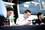 Three men sitting at a table signing autographs during an outdoor event with racing team branding.