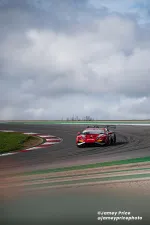 Red race car speeding around a track bend under a cloudy sky during a motorsport event.