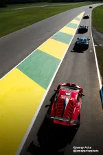 Red race car number 25 leads a pack of sports cars on a racetrack with yellow and green curbs under bright sunlight.