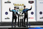 Two race car drivers in blue and black suits celebrating with a trophy at Daytona International Speedway podium.