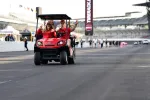 Group of people riding a red golf cart on a racetrack with grandstands in the background