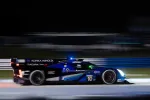 Blue and black Acura Konica Minolta race car speeding on track at night with motion blur and headlights on