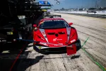 Red race car in pit lane during motorsport event with crew and equipment nearby under clear sky.