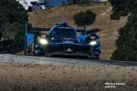Blue Acura Konica Minolta race car with headlights on racing on a track under clear sky