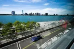 Blue race car speeding on a city waterfront track with urban skyline and water in the background under clear sky