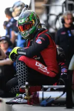 Race car driver in colorful helmet and red racing suit crouching in pit lane with team members in background.