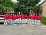 Group of 13 painters in red shirts and white pants posing outdoors near a tree and parked cars.