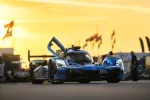 Blue Acura race car with open door on racetrack at sunset with flags and people in background