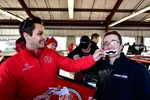 Men in racing jackets smiling and playing with fake mustaches inside a garage at a motorsport event.