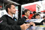 Race car drivers wearing black jackets sign autographs for fans at a motorsport event booth.