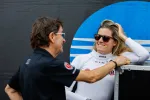 Female race car driver in white suit and sunglasses chatting with a man in black shirt at a racing event.