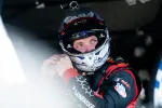 Race car driver wearing helmet and racing suit adjusts gear before a race in the pit area.