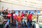 Diverse group of people posing inside racing tent between two race cars, some wearing racing gear and waving.