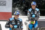 Two Acura racecar drivers celebrate with champagne spray on the Road America podium wearing blue and black racing suits.