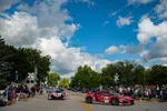 Red and black race cars driving through a street lined with spectators under a blue sky with clouds.