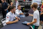 Race car drivers sign autographs for fans at a motorsport event with tires and team members in the background