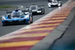 Blue Acura race car leads two other prototype cars on a curving race track with red and yellow curbs.