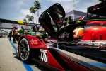 Red and black race car number 40 with open door and driver wearing orange helmet on racetrack pit lane.