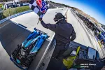 Race official waves checkered flag as blue Acura race car crosses the finish line on a sunny day.