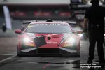 Red Lamborghini race car with headlights on in pit lane during a motorsport event at dusk.