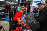 Female racer signs a large racing tire covered with signatures at Acura motorsports event with fans.