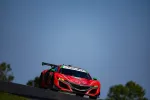 Red Acura race car speeding on track under clear blue sky with green trees in background.