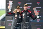 Two race car drivers in black suits and Pirelli hats celebrate holding trophies on a podium with event branding behind them.