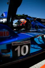 Blue racing car number 10 with driver wearing helmet inside cockpit under bright daylight.