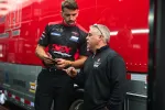 Two men in racing team uniforms discussing near a red and gray race truck, one holding a tablet.