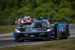 Blue and black Acura Konica Minolta race car on track with blurred cars and green trees in background