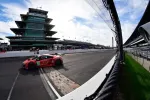 Red race car crossing the iconic bricks at Indianapolis Motor Speedway under a partly cloudy sky.