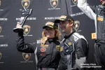 Two race car drivers in black racing suits and Pirelli hats celebrating with trophies on a Lamborghini podium.