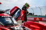 Race car driver in red and black suit with green helmet stands beside open door of Acura race car on track.