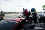 Race car driver climbing into a red racing car with another driver and crew at a racetrack pit stop.