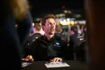 Man in black Konica Minolta jacket speaking at a table with blurred crowd and lights in the background at night.