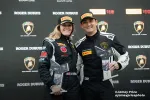 Two race car drivers in black racing suits and Pirelli hats holding trophies smiling in front of Lamborghini and Roger Dubuis logos.