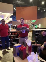 Happy man in red shirt holding a wooden gift plaque inside a festive bowling alley with Christmas decorations.