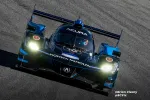 Black and blue Acura prototype race car with headlights on racing on asphalt track.