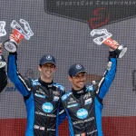 Two race car drivers in blue Konica Minolta suits celebrating with trophies on a podium after winning third place.
