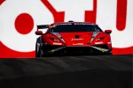 Red and black race car speeding on track with bold red and white background sign visible behind.