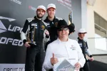 Race car drivers and a man in a cowboy hat hold trophies and celebrate on the podium after a motorsport event.
