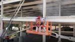Worker in safety gear sandblasting or cleaning metal beams on building structure from elevated orange platform.