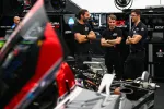 Three men in black shirts discuss near a partially disassembled race car in a professional racing garage.